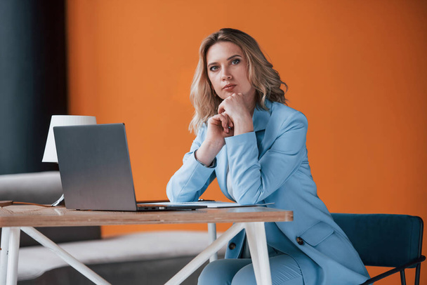 Keep talking, I listen to you. Businesswoman with curly blonde hair indoors in room with orange colored wall and wooden table. - Фото, изображение