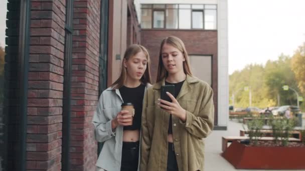 Seguimiento de tiro de alegres hermanas gemelas jóvenes caminando por la calle y charlando mientras se mira el teléfono móvil - Metraje, vídeo