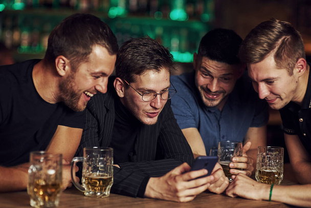 Funny content on smartphone. Three friends resting in the pub with beer in hands. Having conversation. - Zdjęcie, obraz