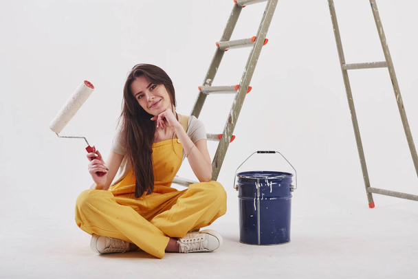 Awesome work with painting the walls. Beautiful brunette in yellow uniform and blue colored bucket sits near the ladder. - Photo, image