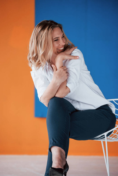 Nice joke. Businesswoman with curly blonde hair sitting on white chair indoors in room with orange and blue colored wall. - Фото, изображение