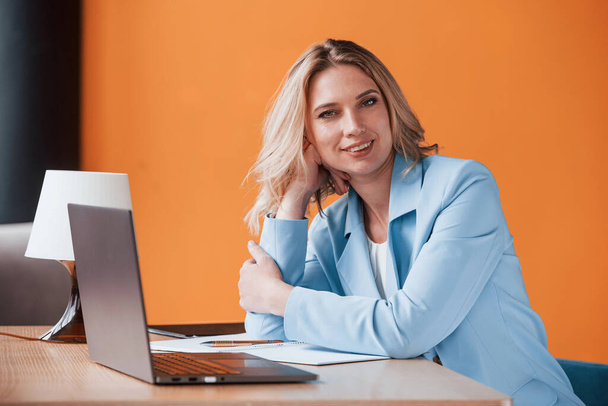 Welcome to my workplace. Businesswoman with curly blonde hair indoors in room with orange colored wall and wooden table. - Photo, Image