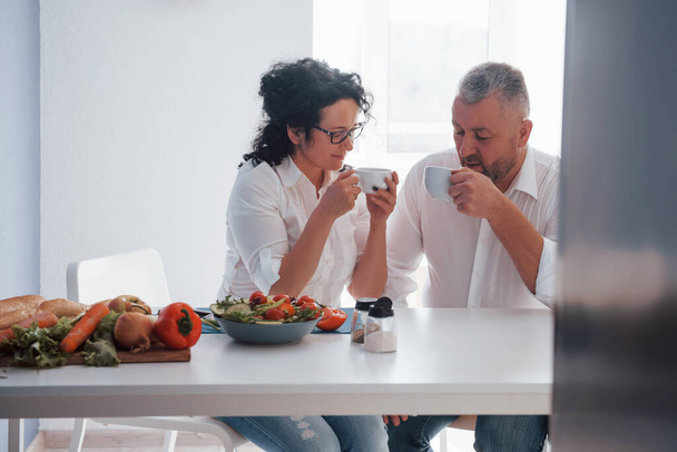 Calm time spending. Senior man and his wife in white shirt have romantic dinner on the kitchen. - Foto, afbeelding