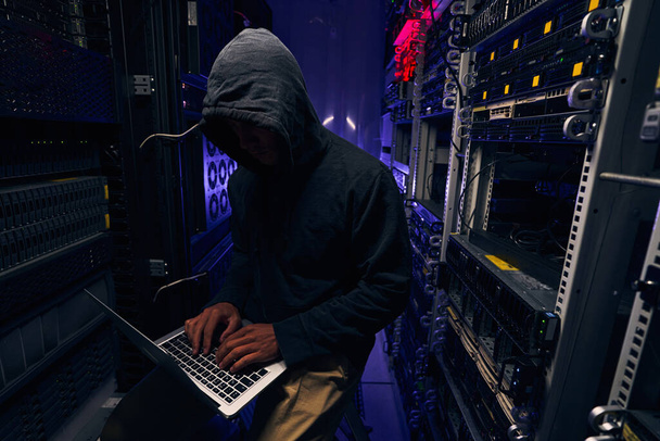 Focused hacker typing on laptop in server room - Photo, image