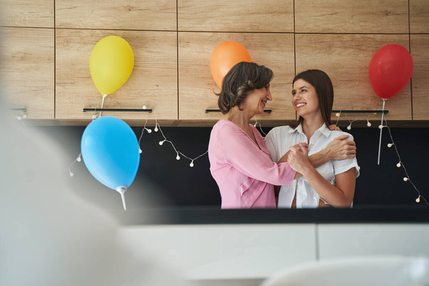 Smiling adult woman and young female person hugging in kitchen - Foto, immagini