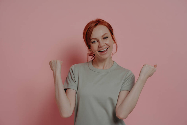 Overjoyed red-haired woman celebrating victory, doing winner gesture on pink background - Fotografie, Obrázek