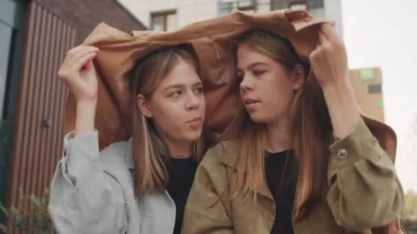 Niski kąt ujęcia szczęśliwych młodych bliźniaczek ukrywających się pod płaszczem i śmiejących się siedząc na zewnątrz - Materiał filmowy, wideo