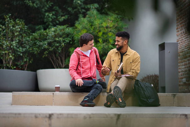 Onnellinen nuori mies Downin oireyhtymä ja mentorointi ystävä istuu ja puhuu ulkona - Valokuva, kuva