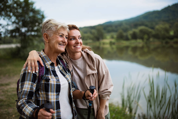 Happy senior mother hiker embracing with adult daughter when looking at lake outdoors in nature - Фото, изображение