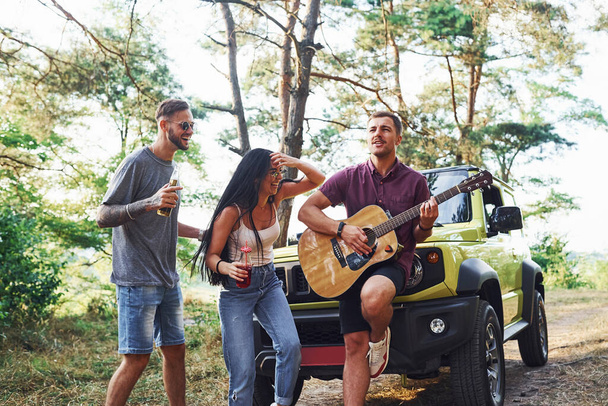 Singing songs together. Friends have nice weekend outdoors near theirs green car with acoustic guitar. - Fotoğraf, Görsel
