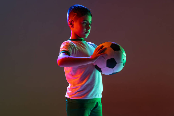Cropped portrait of boy, child, football player training isolated over gradient background in neon light. - Zdjęcie, obraz