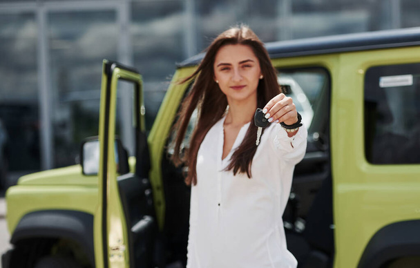 Positive face expression. Holds keys of her new vehicle. Young woman in white official clothes stands in front of green automobile outdoors. - Fotó, kép