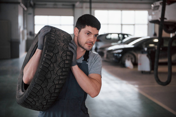 Short haired unshaved man. Mechanic holding a tire at the repair garage. Replacement of winter and summer tires. - Foto, imagen