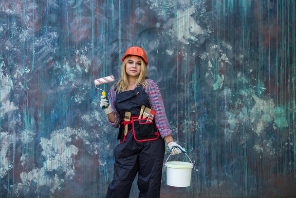 jolie femme en uniforme et casque tenant peinture et rouleau se préparant à rénover la maison. Travailleur de la construction. - Photo, image