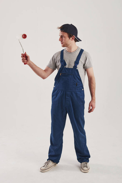 Roller for painting walls. Man in blue uniform stands against white background in the studio. - Foto, afbeelding