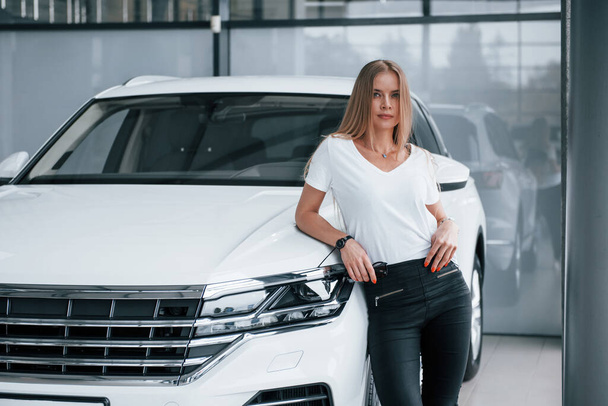 Looking at the camera. Girl and modern car in the salon. At daytime indoors. Buying new vehicle. - Foto, Imagem