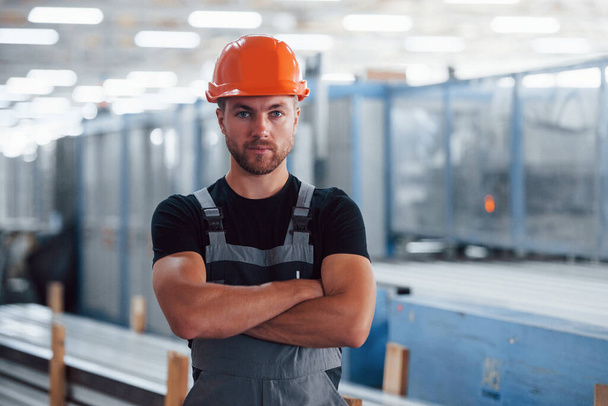 Stands with arms crossed. Portrait of male industrial worker indoors in factory. Young technician with orange hard hat. - Foto, Imagen