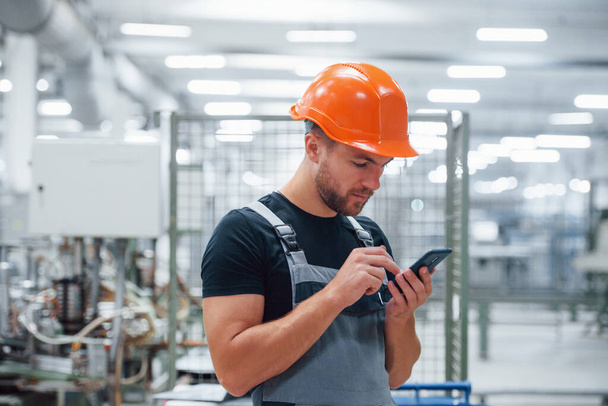Smartphone in hands. Industrial worker indoors in factory. Young technician with orange hard hat. - Photo, image