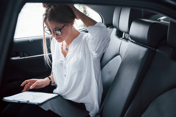 Working on the road. Smart businesswoman sits at backseat of the luxury car with black interior. - Foto, afbeelding