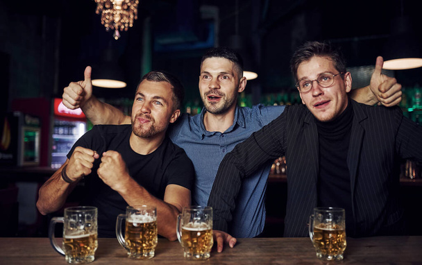 Drie sportfans in een bar die voetbal kijken. Met bier in handen. - Foto, afbeelding