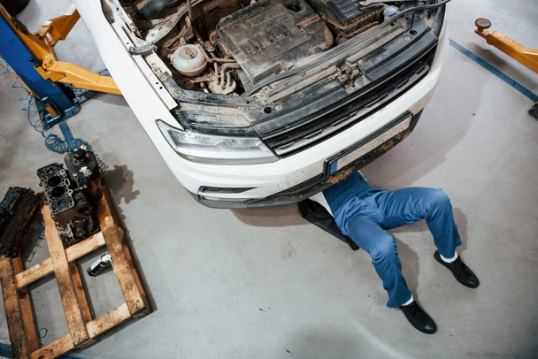 Under the car. Employee in the blue colored uniform works in the automobile salon. - 写真・画像