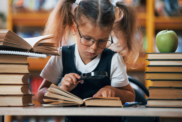 Front view. Cute little girl with pigtails is in the library. Apple on the books. - Fotoğraf, Görsel