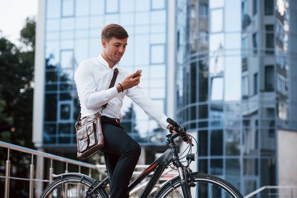Brown bag and pink phone. Businessman in formal clothes with black bicycle is in the city. - Foto, imagen
