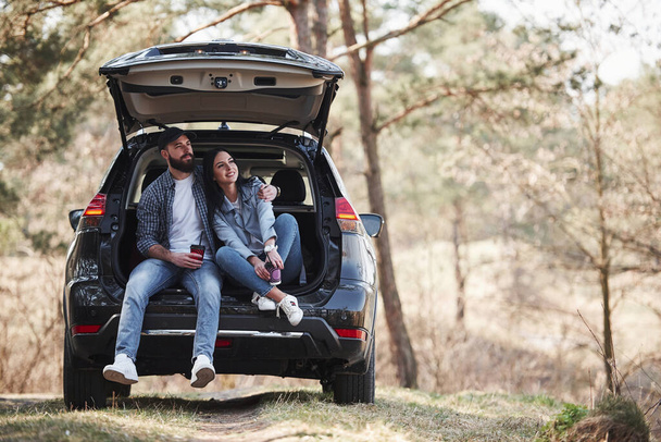 Having a break. Sitting on rear part of automobile. Enjoying the nature. Couple have arrived to the forest on their brand new black car. - Foto, immagini