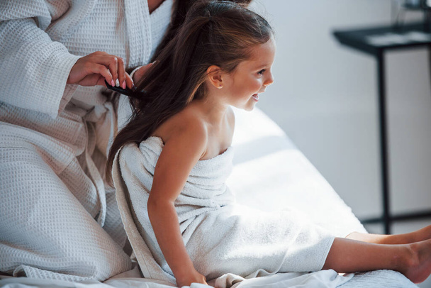 Making hairstyle by using comb. Young mother with her daugher have beauty day indoors in white room. - Foto, Imagem