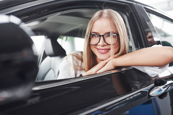 Nice reflection. Beautiful blonde girl sitting in the new car with modern black interior. - Zdjęcie, obraz