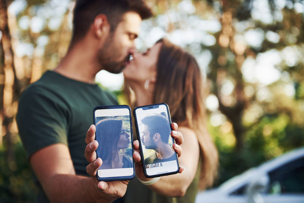 Holding two smartphones with photos of them. Beautiful young couple have a good time in the forest at daytime. - Φωτογραφία, εικόνα
