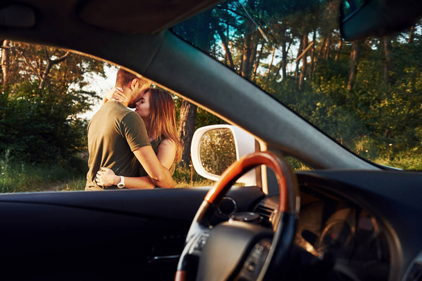 Kilátás az autó belsejéből. Kormánykerék, oldalsó tükör. Gyönyörű fiatal pár szórakozik az erdőben napközben.. - Fotó, kép