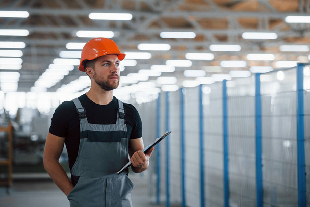 Stands in the corridor with documents in hands. Industrial worker indoors in factory. Young technician with orange hard hat. - Foto, Bild