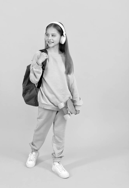 щаслива дівчинка-підліток з шкільним рюкзаком і дошкою копійки слухати музику в навушниках, шкільний час
. - Фото, зображення