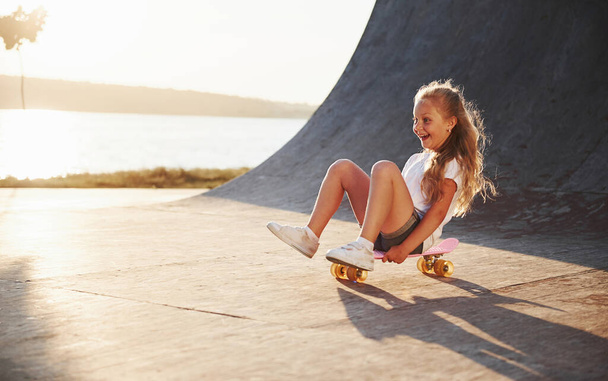 Sits on the skate. Sunny day. Kid have fun at the ramp. Cheerful little girl. - Фото, изображение