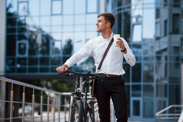 Holding cup with fresh drink. Businessman in formal clothes with black bicycle is in the city. - Zdjęcie, obraz