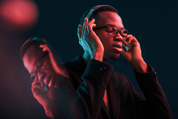 Enjoying listening music in headphones. In glasses. Futuristic neon lighting. Young african american man in the studio. - Photo, Image