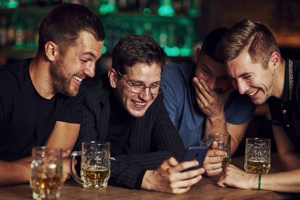 Funny content on smartphone. Three friends resting in the pub with beer in hands. Having conversation. - Foto, Bild