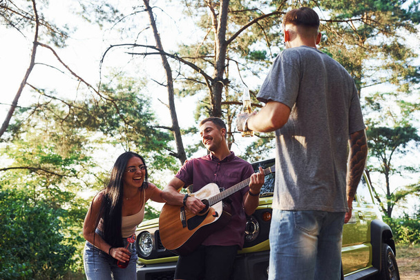 Singing songs together. Friends have nice weekend outdoors near theirs green car with acoustic guitar. - Zdjęcie, obraz
