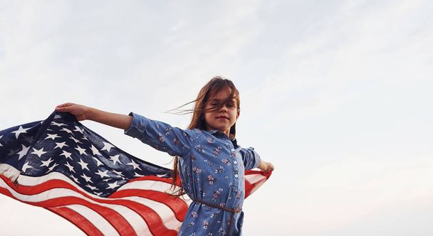 Patriotic female kid with American Flag in hands. Against cloudy sky. - 写真・画像