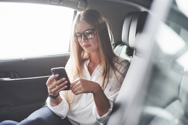 Using smartphone. Beautiful blonde girl sitting in the new car with modern black interior. - Foto, Imagen