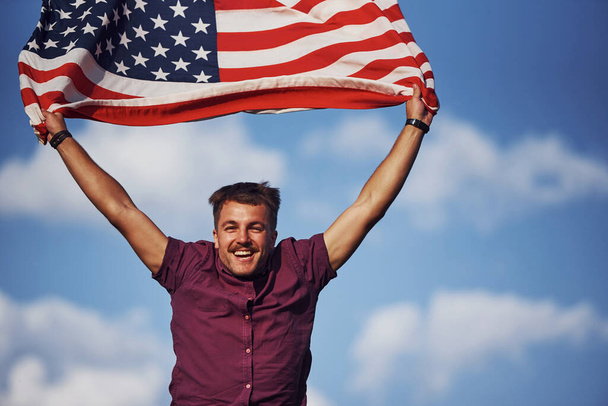 Patriotic happy man waving American Flag against cloudy blue sky. - Фото, зображення