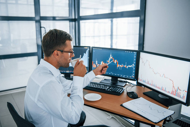 Holds cup of drink. Analysing information. Young businessman in formal clothes is in office with multiple screens. Conception of exchange and money. - Photo, Image