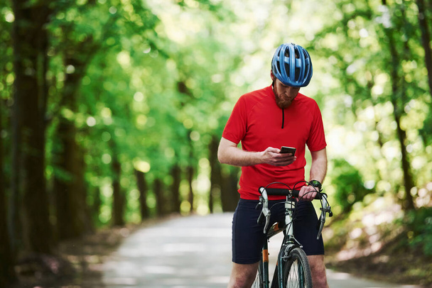Holding smartphone. Cyclist on a bike is on the asphalt road in the forest at sunny day. - 写真・画像
