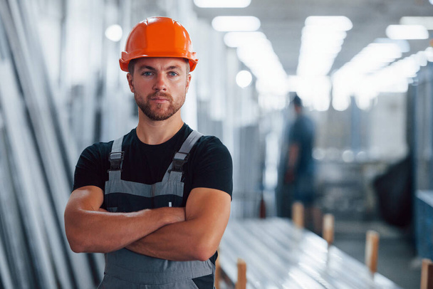 Stands with arms crossed. Portrait of male industrial worker indoors in factory. Young technician with orange hard hat. - Foto, Bild