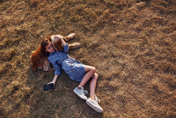 Top view. Lying on the grass with black phone. Cute little girl have a walk with her dog outdoors at sunny day. - Фото, зображення