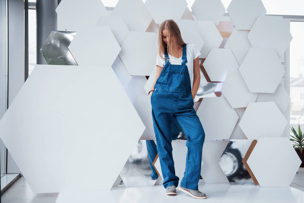 Девушка в синей форме стоит напротив абстрактной стены с белыми кусочками поверхности в форме шестиугольника в автосалоне. - Фото, изображение