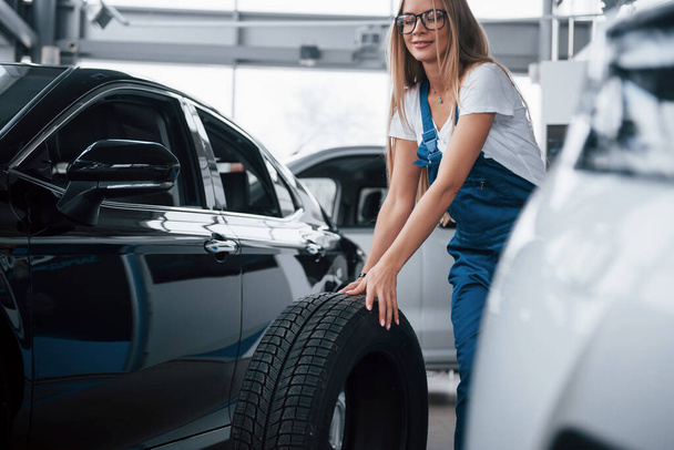 Luxury black vehicle. Woman walks with brand new wheel to the car. Conception of repair. - Photo, image