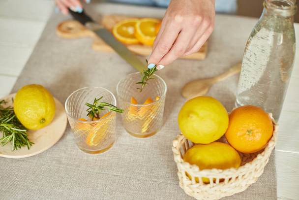 Female hand put rosemary in glasses, Woman preparing, making citrus and rosemary fresh lemonade in glass on a white table at home, summer drink, detox healthy water. - Photo, Image