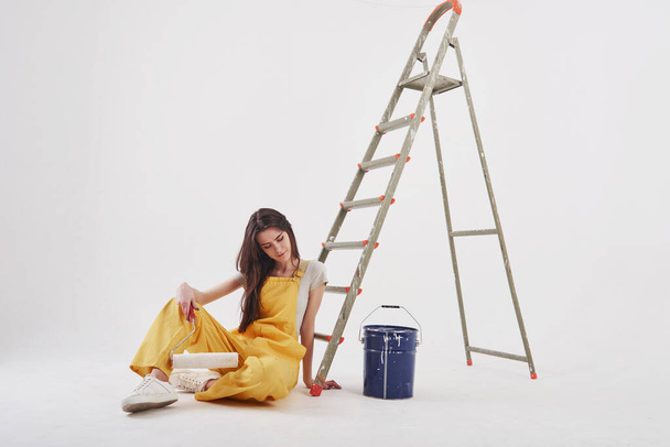 The hard work gave results. Beautiful brunette in yellow uniform and blue colored bucket sits near the ladder. - Фото, изображение
